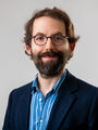 Mitarbeiterfoto Richard Schumm: Lächelnder Mann mit dunklen Haaren, Brille und Bart mit mit dunklem Sakko und gestreiftem Hemd