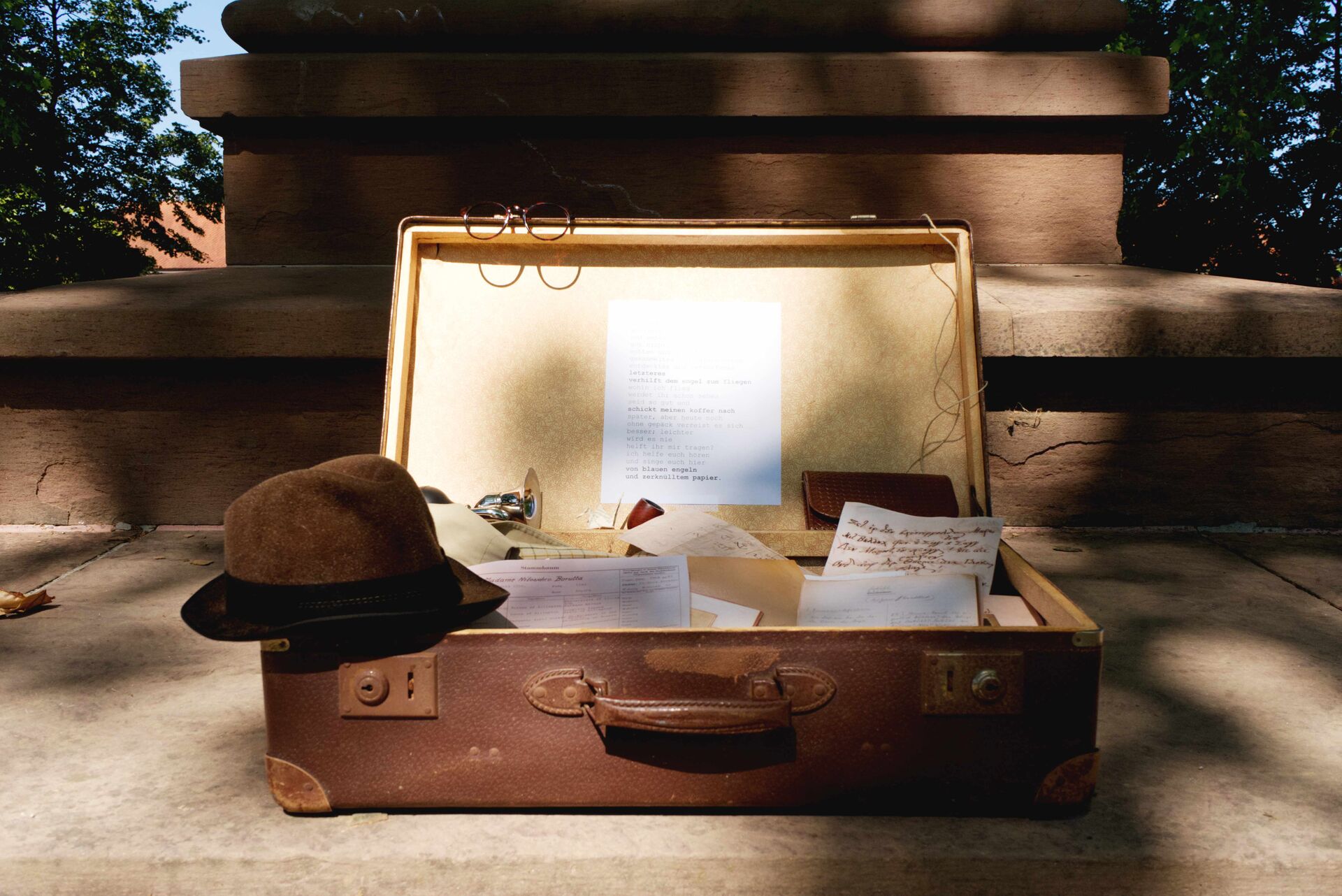 Symbolbild: Geöffneter Koffer mit Papieren, einem Hut auf dem linken Rand und einer Brille auf der geöffneten Klappe 